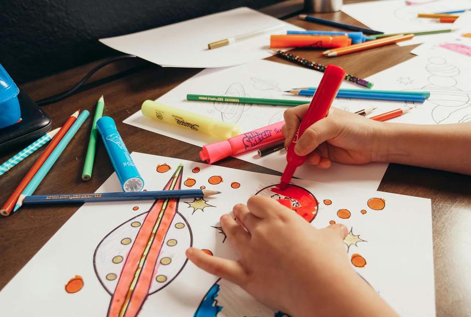 Paper, color pencils and markers on table with child's hand holding a color marker drawing space ships