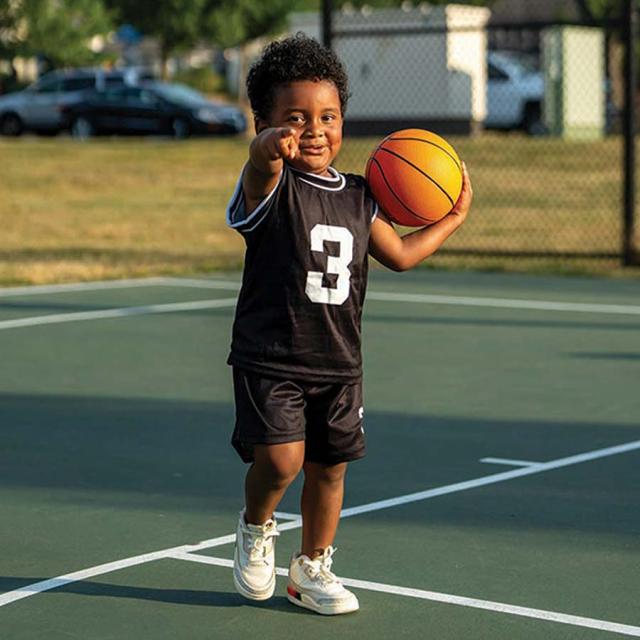 Young 4 year old boy on the basketball course holding a basketball and pointing to the camera laughing ready to play some ball.